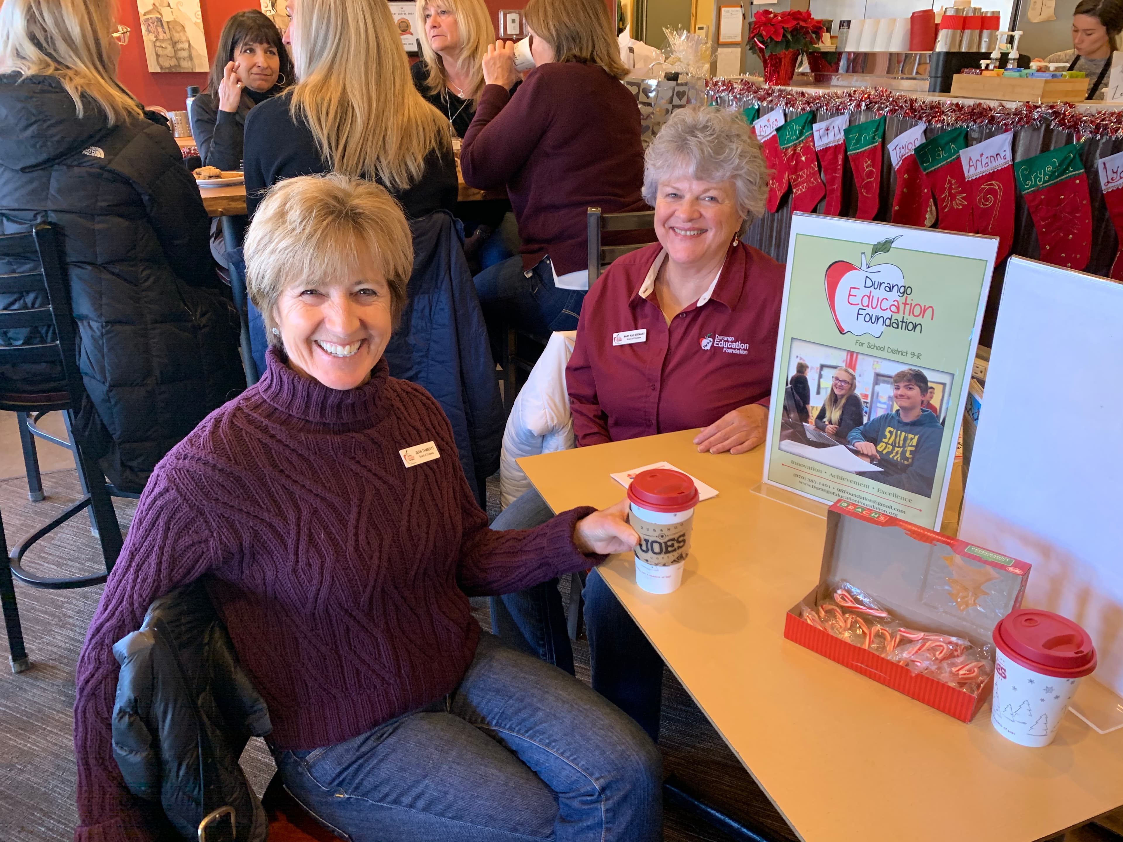 Two DEF trustees, one in a maroon DEF polo, at a community table with a Durango Education Foundation sign